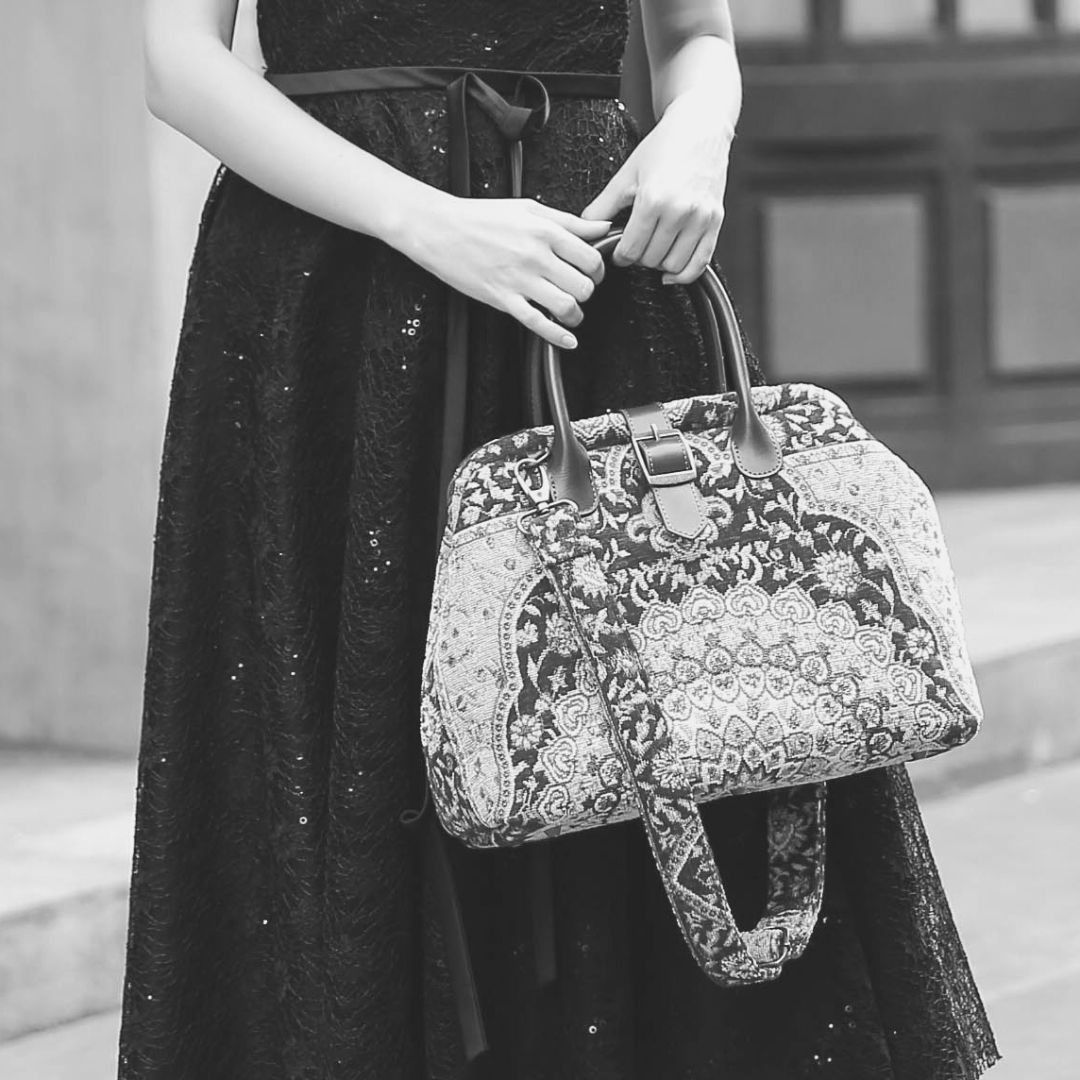 Woman wearing a black sequined dress carrying a vintage-inspired carpet handbag with leather handles and shoulder strap.