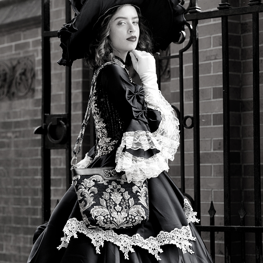 Woman in a Victorian-inspired black dress with lace details and a wide-brimmed hat, carrying a floral tapestry shoulder bag with a butterfly clasp while posing beside an iron gate.