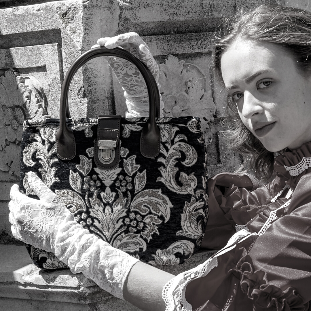 Victorian-style woman in a lace-trimmed dress and gloves holding a black damask tapestry handbag with leather handles and clasp closure.